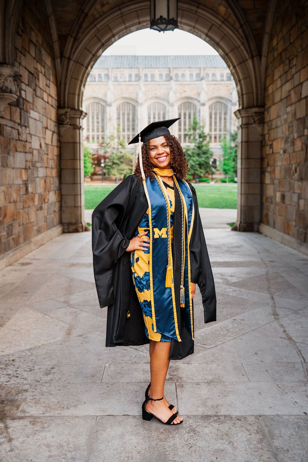 Graduate in cap and gown poses under stone archway during commencement photo session at university campus.