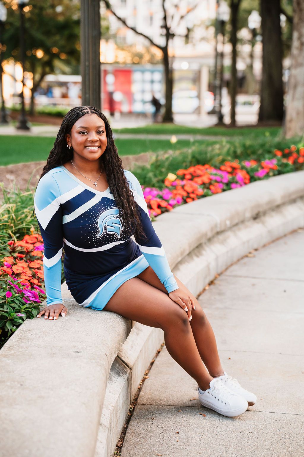 Cheerleader in navy and light blue uniform poses on curved concrete wall near colorful flower garden.
