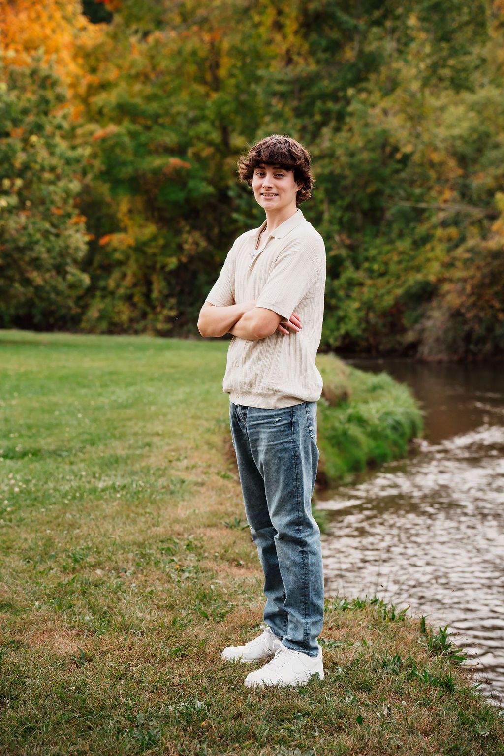 Teen boy with curly hair stands arms crossed by a creek in autumn, wearing jeans and a cream sweater.