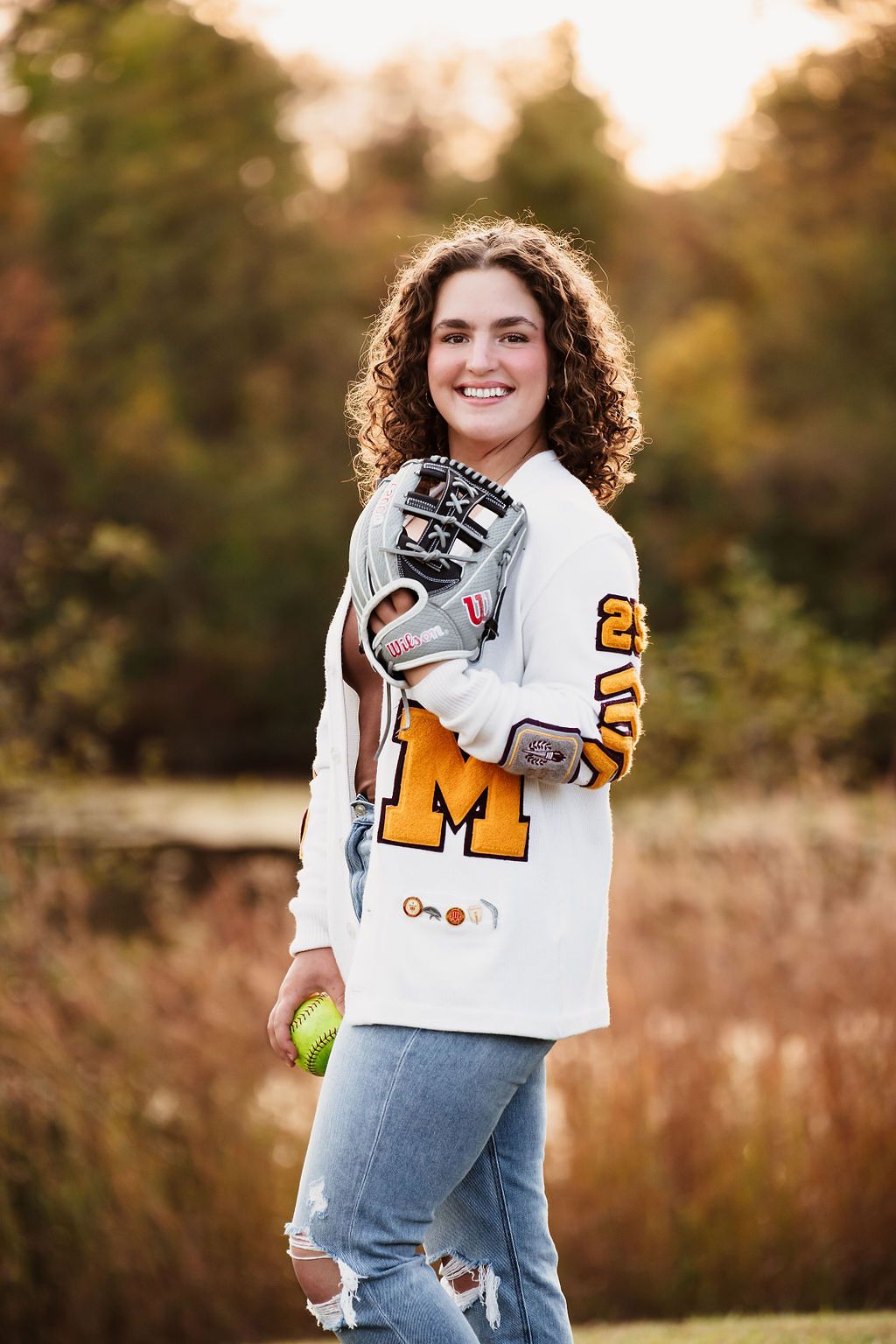 Smiling teen girl in white varsity jacket holds softball glove and ball outdoors in autumn setting.