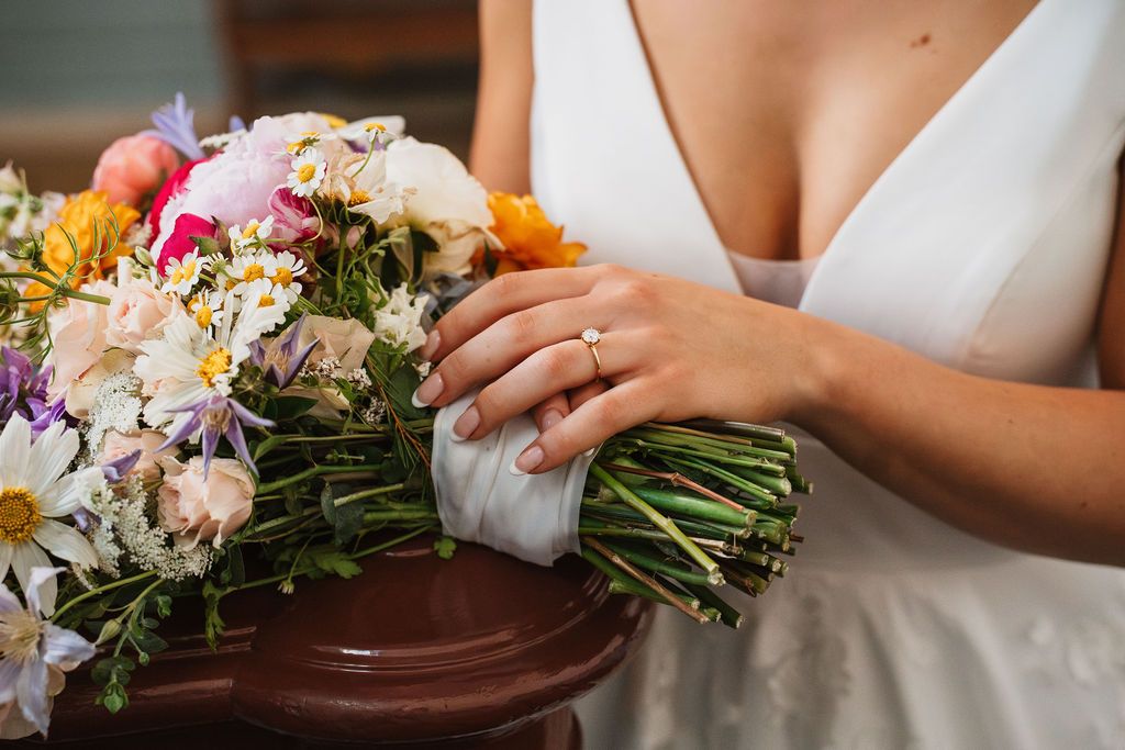 Colorful bridal bouquet with mixed flowers held against white wedding dress.