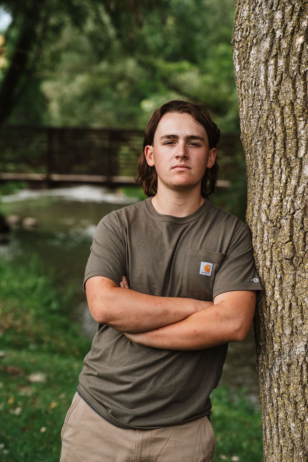 Person wearing a gray Carhartt t-shirt poses against a tree trunk in an outdoor setting with greenery in background.