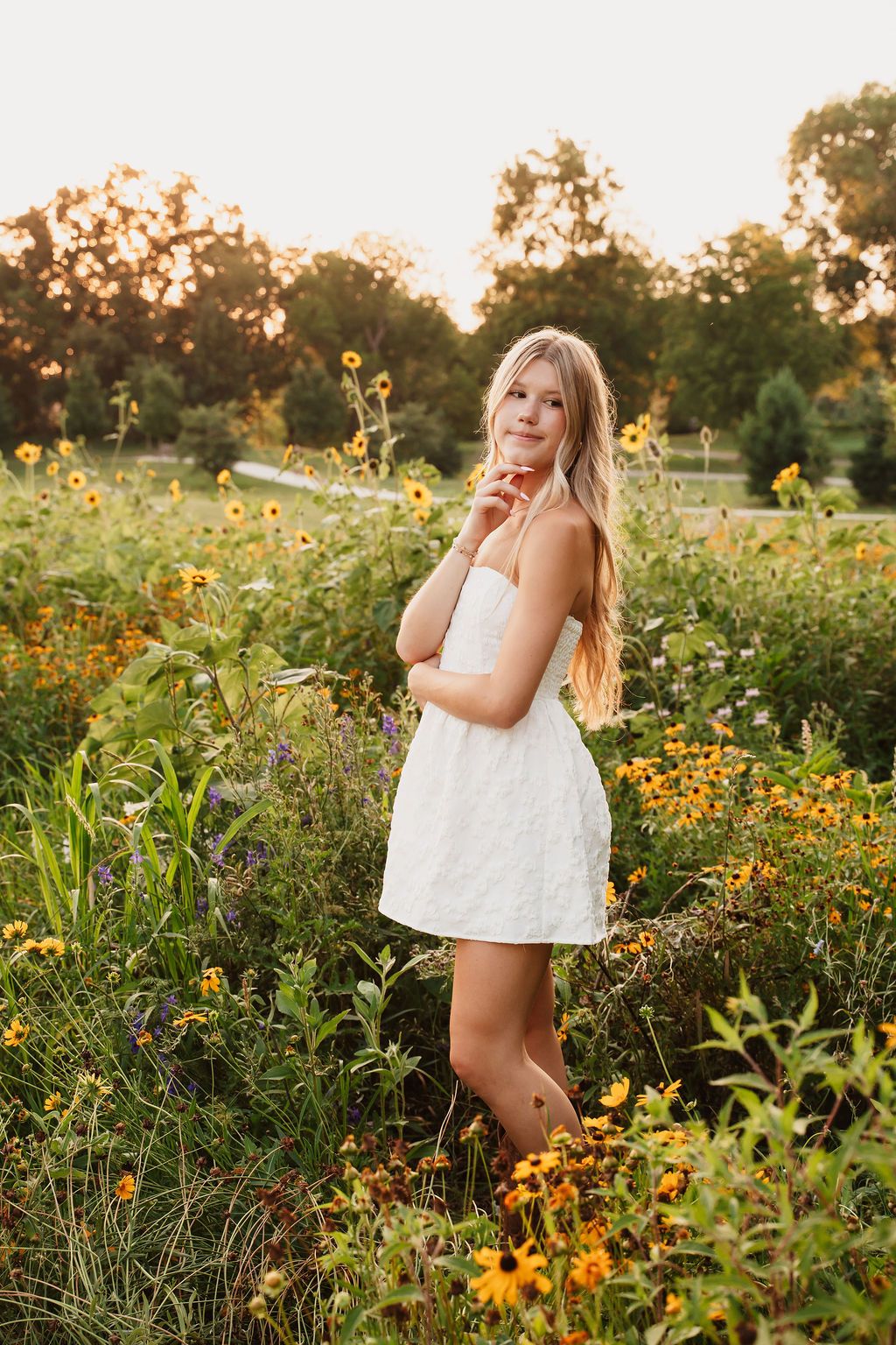 Young blonde woman in white dress standing in a vibrant wildflower field at golden hour sunset.