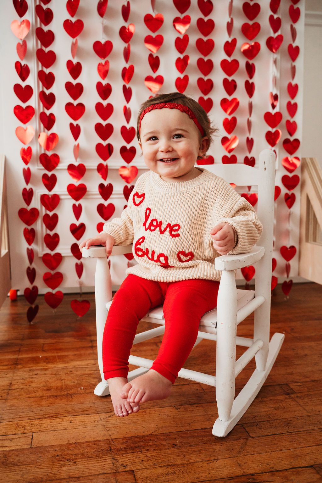 Smiling baby in cream “love bug” sweater and red leggings sits in white rocking chair with Valentine’s Day heart backdrop.