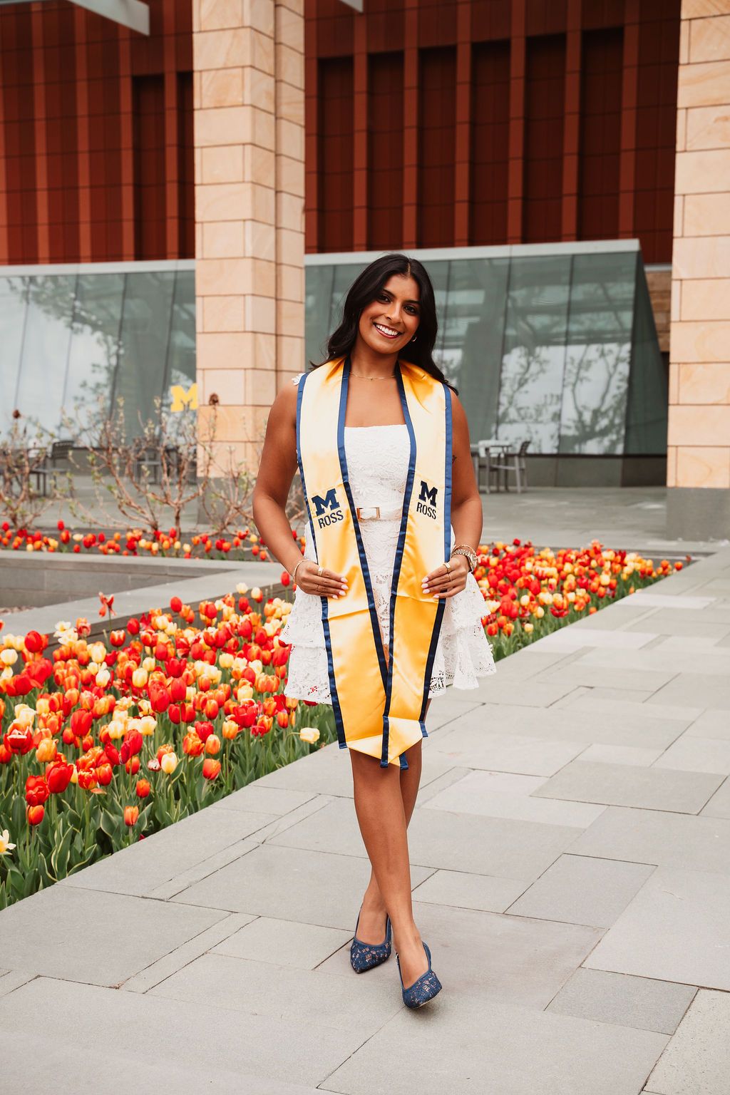 University of Michigan graduate smiling in white dress and gold stole, posing among red and yellow tulips on campus.
