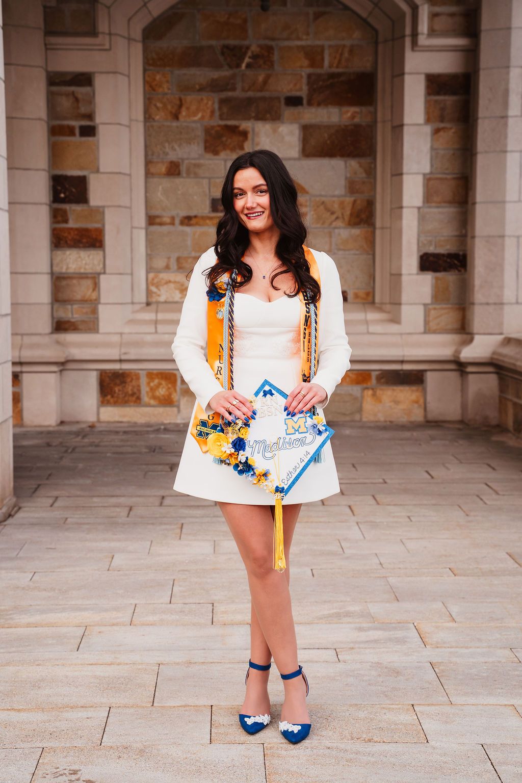 A person in a white dress and yellow cardigan poses playfully in front of a stone building with blue shoes.