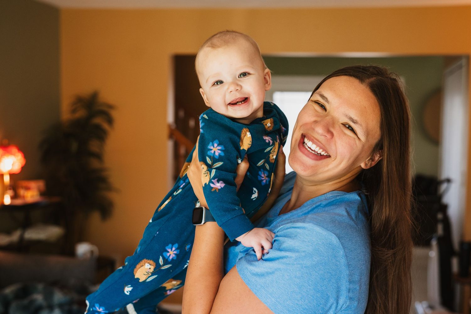 A smiling mother holds her happy baby in a blue floral onesie indoors, sharing a joyful moment together.