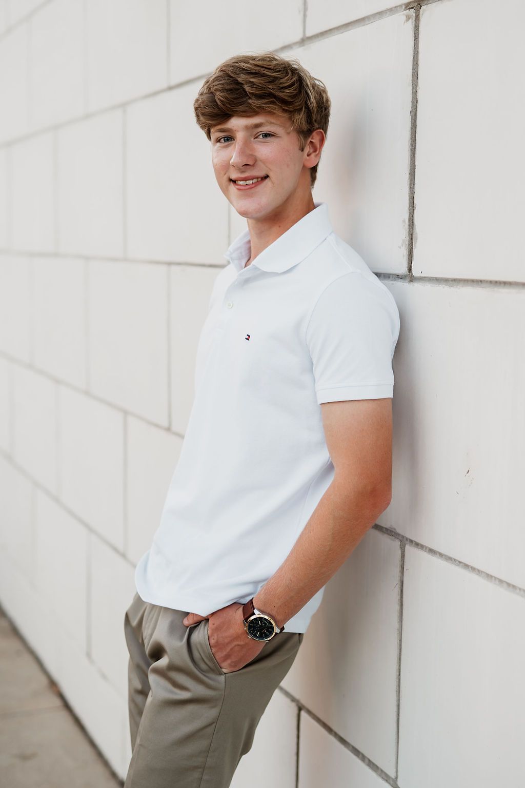 Smiling teen boy in white Tommy Hilfiger polo and khakis leaning against a white brick wall.
