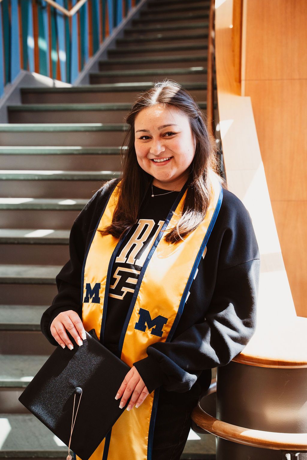 Smiling graduate in University of Michigan sweatshirt and gold stole holds cap on staircase.