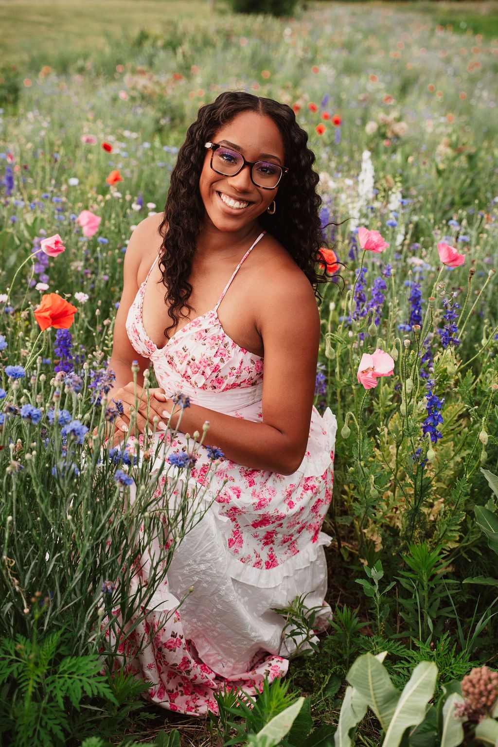 Young woman in floral dress smiling in a vibrant wildflower field with poppies and blue blooms.
