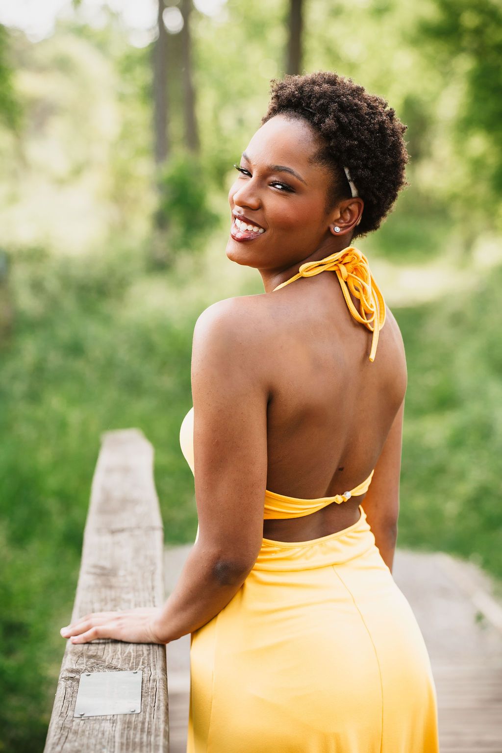 A person in a bright yellow sundress poses on a wooden boardwalk surrounded by lush green foliage on a sunny day.