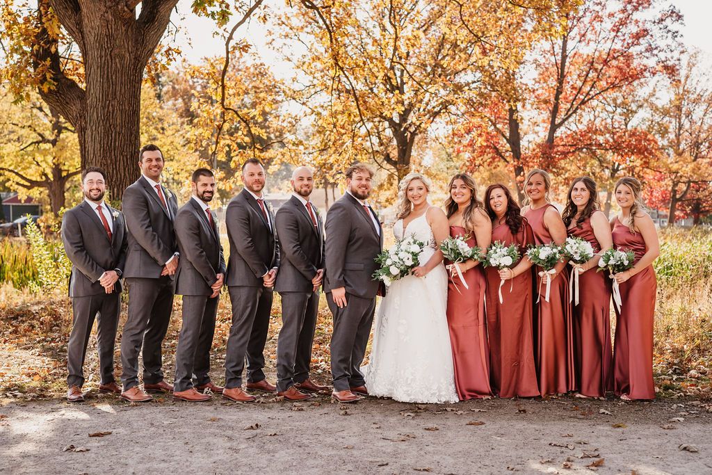 Wedding party poses in front of fall foliage wearing burgundy and gray attire.