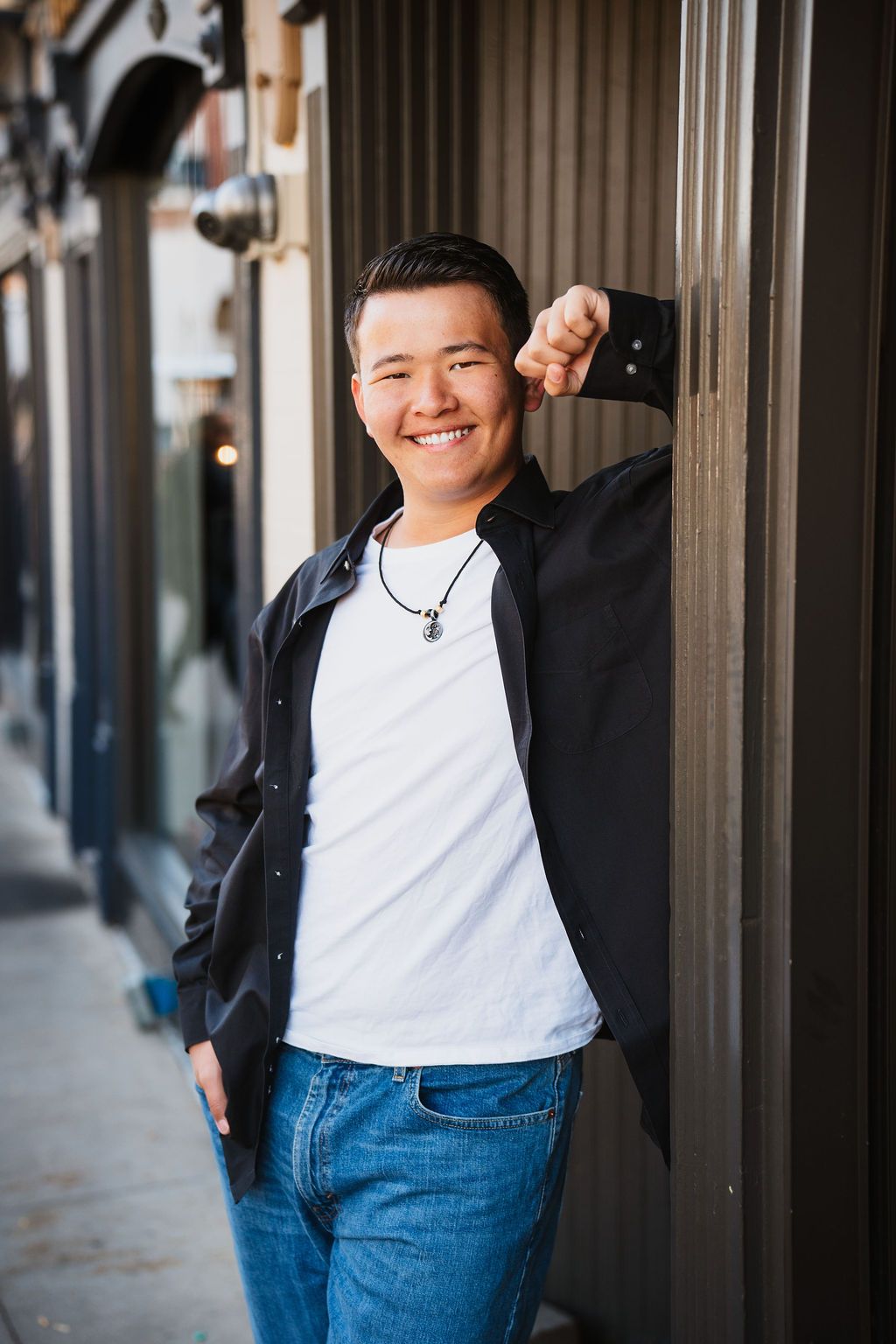 Smiling young man leaning against a dark wall outdoors, wearing a black shirt over a white tee and jeans.