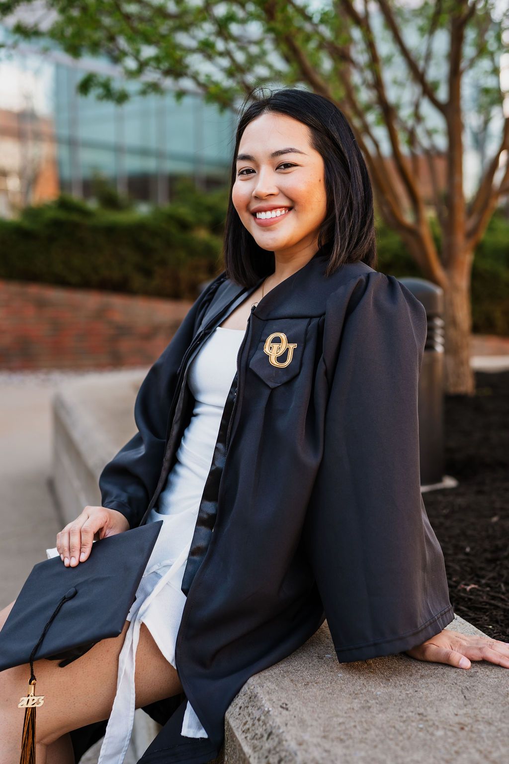 Graduate in black academic regalia sits on concrete ledge near trees and glass building with university emblem.