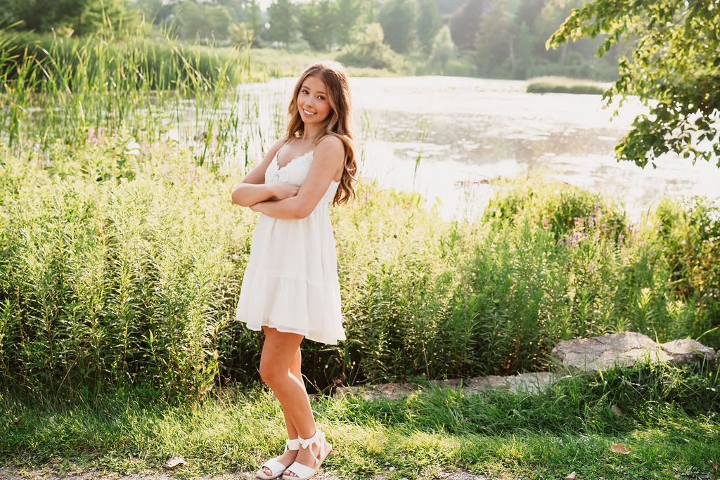 A white lace sundress and sneakers outfit photographed in a sunny lakeside setting with tall grass and trees.