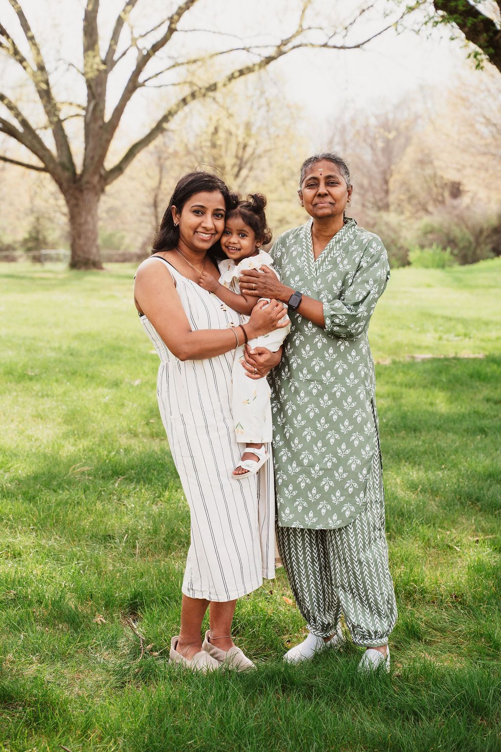 Three generations of South Asian women pose together outdoors in a lush green park, smiling warmly.