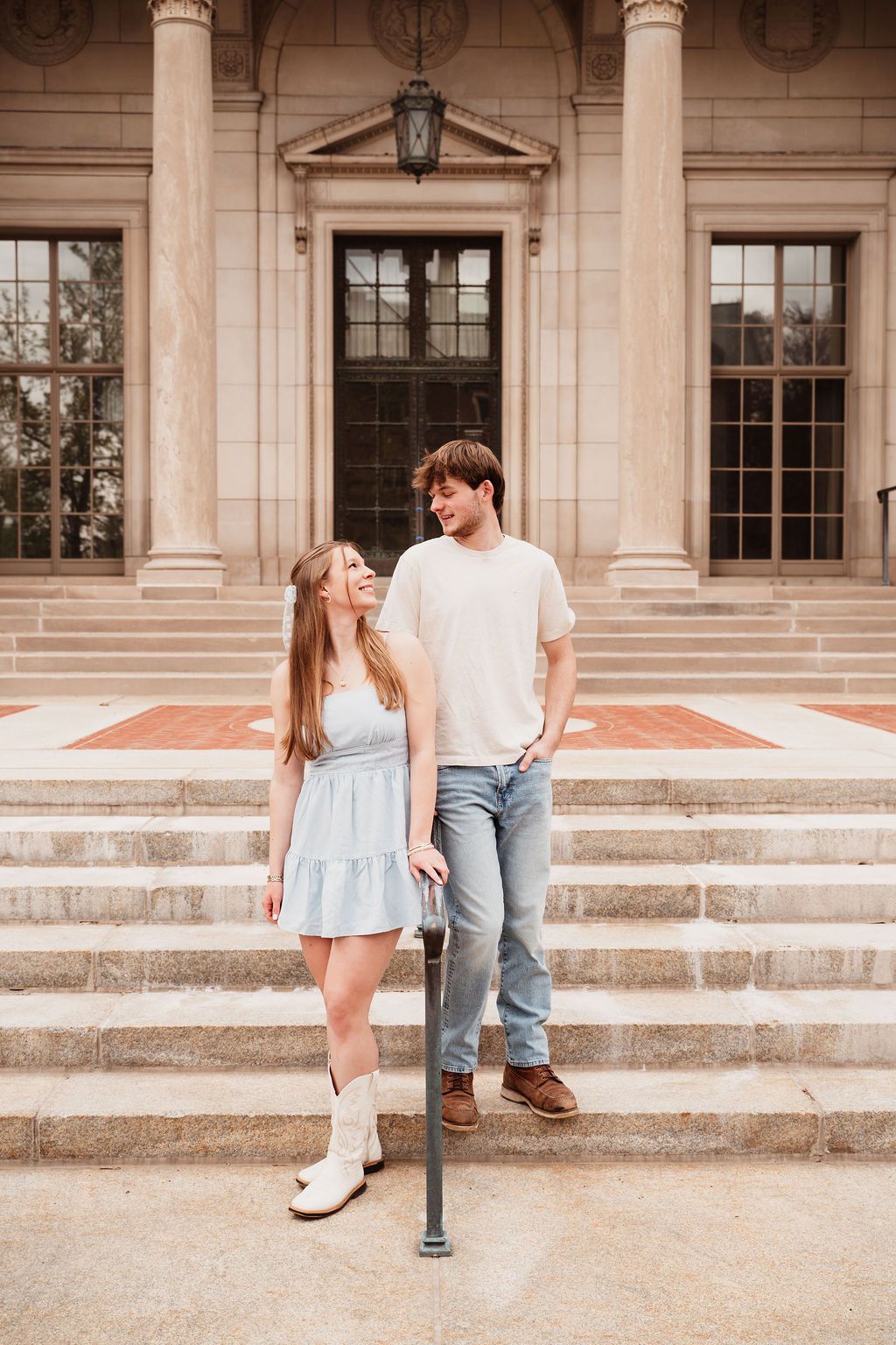 A young couple smiles at each other on stone steps outside a classic building, she in a blue dress and white boots.