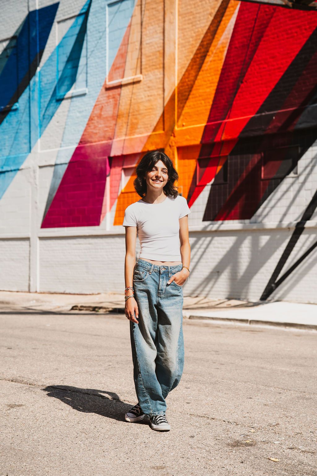 Young woman in white crop top and wide-leg jeans smiling in front of a colorful geometric mural outdoors.