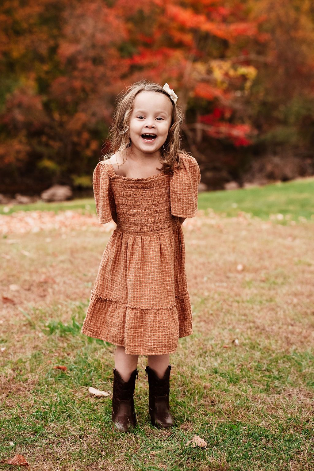A joyful little girl in an orange dress and cowboy boots laughs outdoors among vibrant fall foliage.