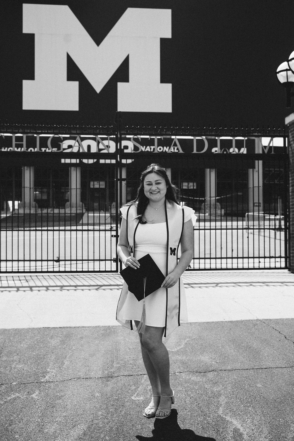 Graduate poses in white dress with cap outside University of Michigan stadium in black and white photo.
