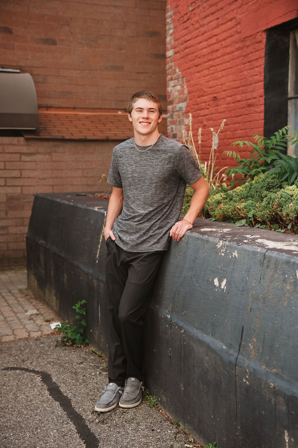 Smiling young man in gray t-shirt and black pants leaning against a wall outdoors near red brick building.