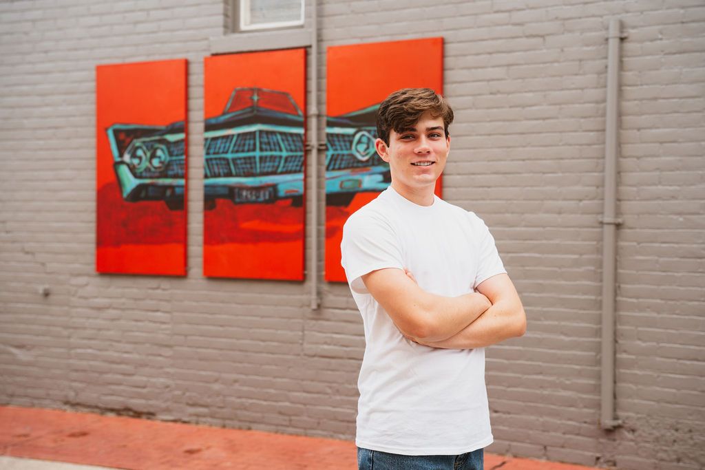 Young man in white t-shirt stands arms crossed before vibrant red vintage car triptych mural on brick wall.