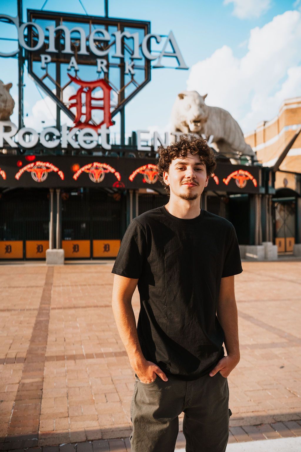 Young man with curly hair in black t-shirt standing outside Comerica Park, home of the Detroit Tigers, on a sunny day.
