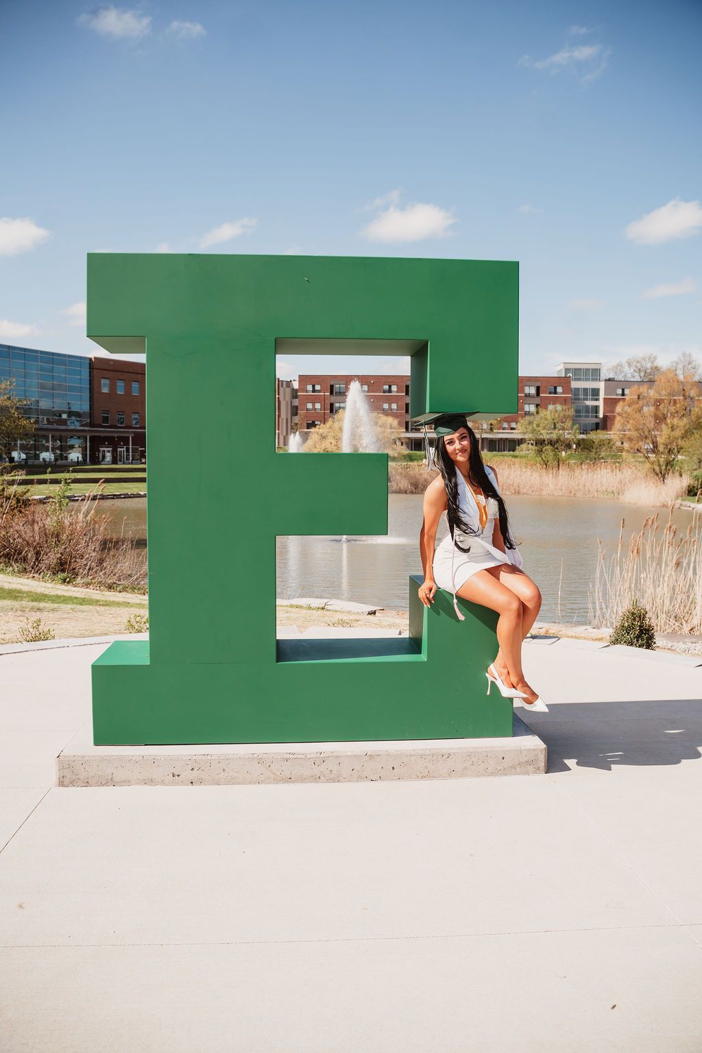 A graduate in cap and gown poses on a large green E letter sculpture at Eastern Michigan University campus.