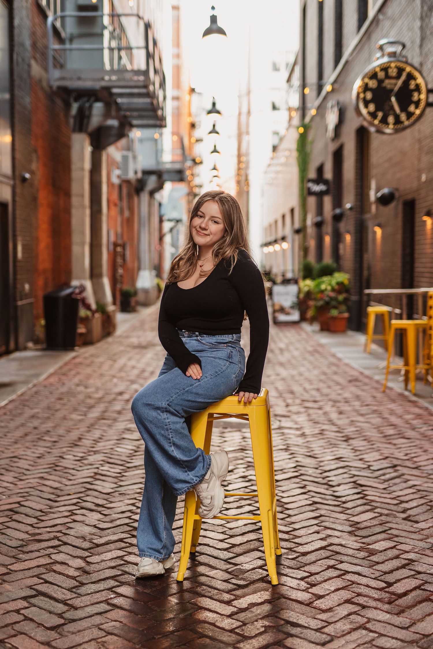 Young woman in black top and wide-leg jeans sits on yellow stool in charming brick alley with string lights.