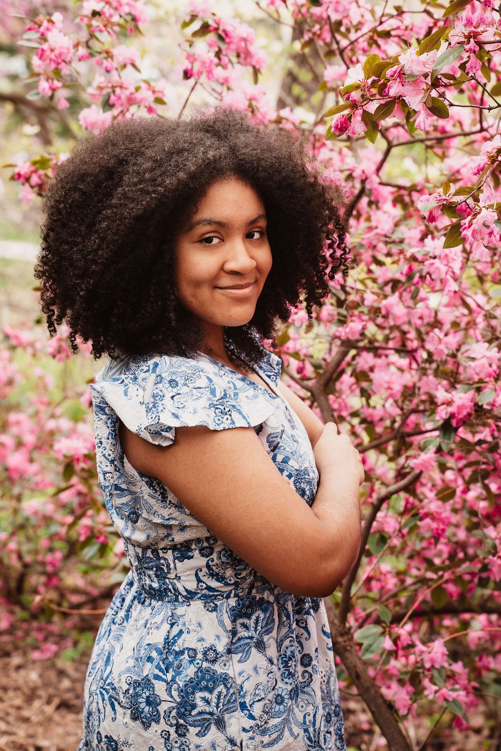 Young Black woman with curly hair in blue floral dress smiling among pink cherry blossom trees in spring.