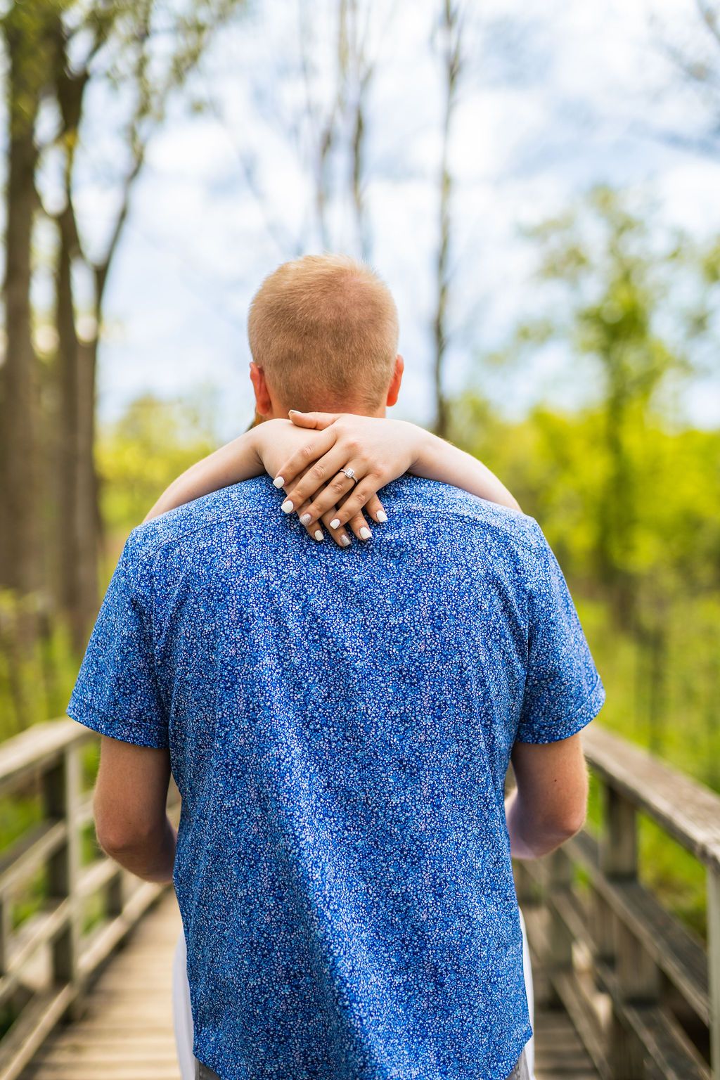 Person in blue speckled t-shirt walking across wooden boardwalk through forest on sunny day.