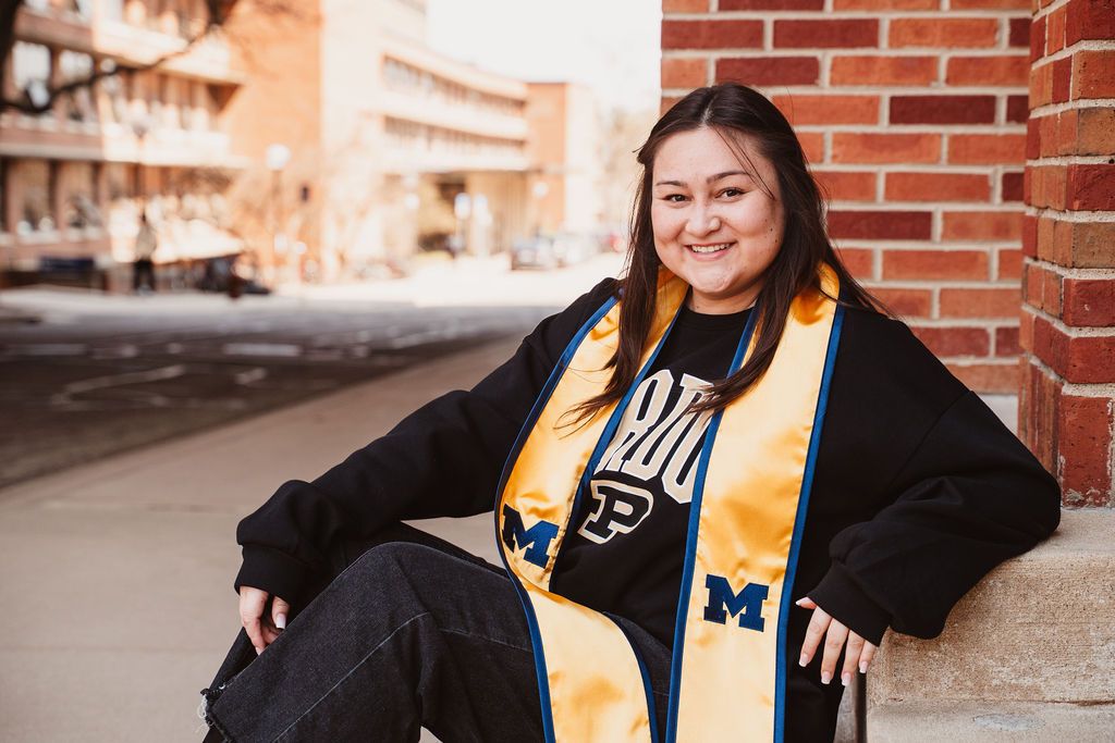 Smiling graduate wearing University of Michigan stole and Purdue shirt leaning against brick wall.