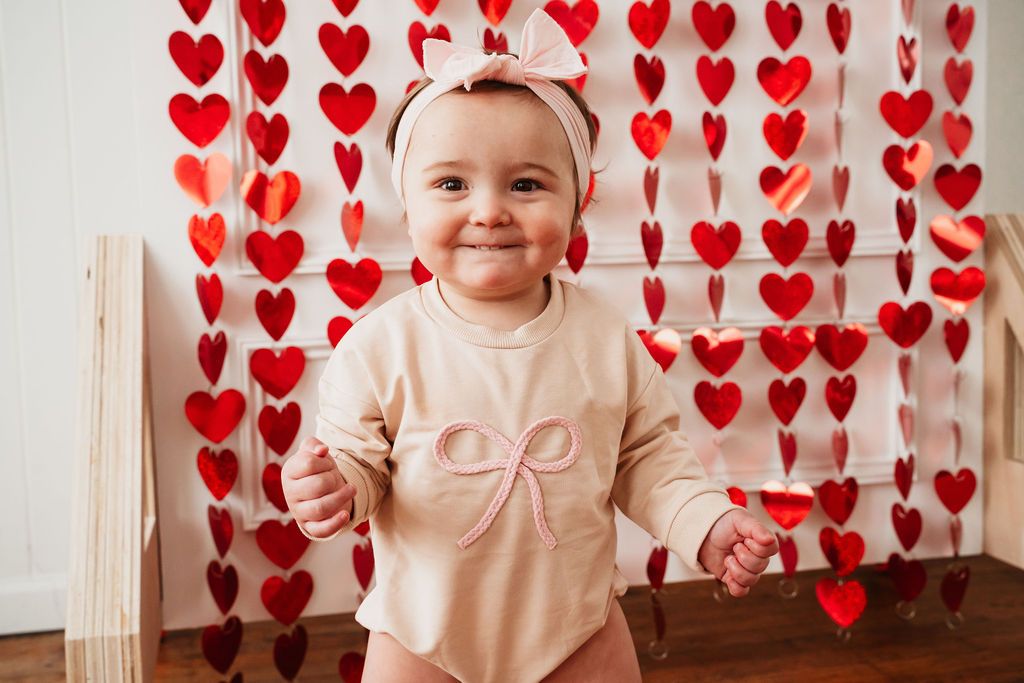 Baby girl in beige bow outfit smiles in front of red heart garland backdrop for Valentine's Day photo.