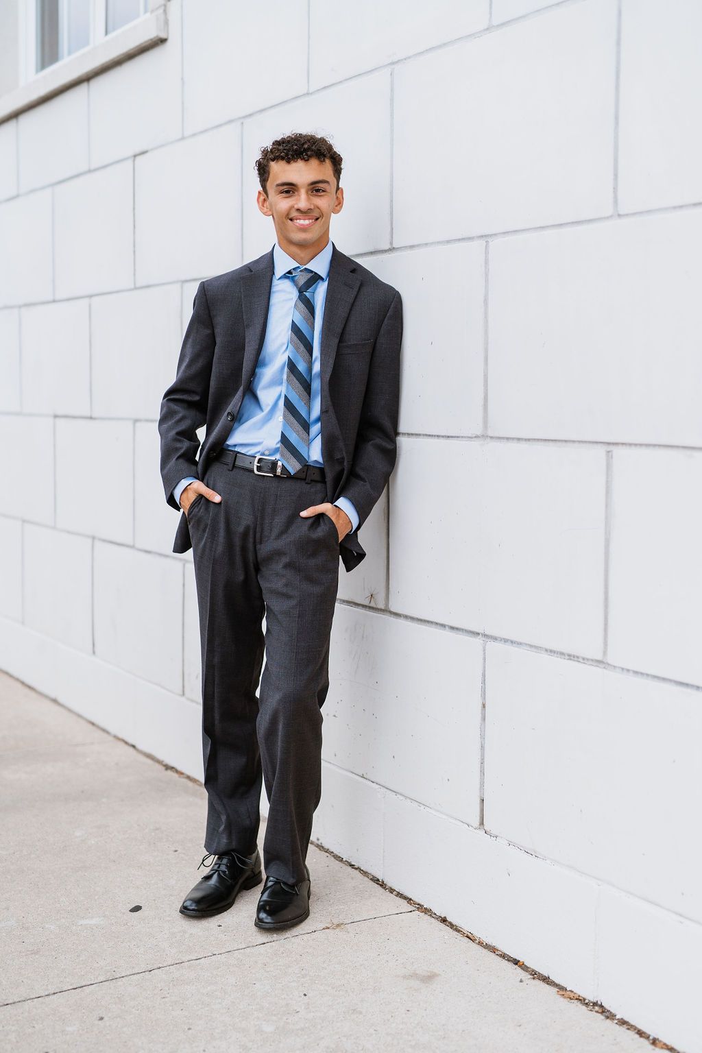 Young man in dark suit, blue striped tie, smiling confidently while leaning against a white wall outdoors.