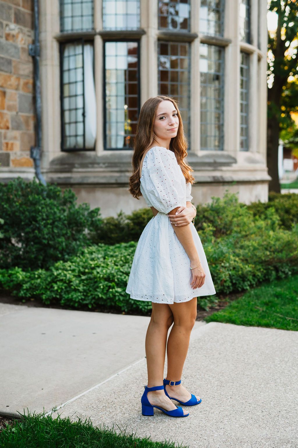 A person in a white lace dress and bright blue heels stands on a gravel path near a historic stone building.