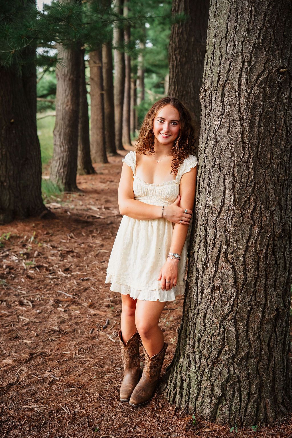 A white lace dress and brown boots create a rustic woodland portrait scene among tall pine trees.