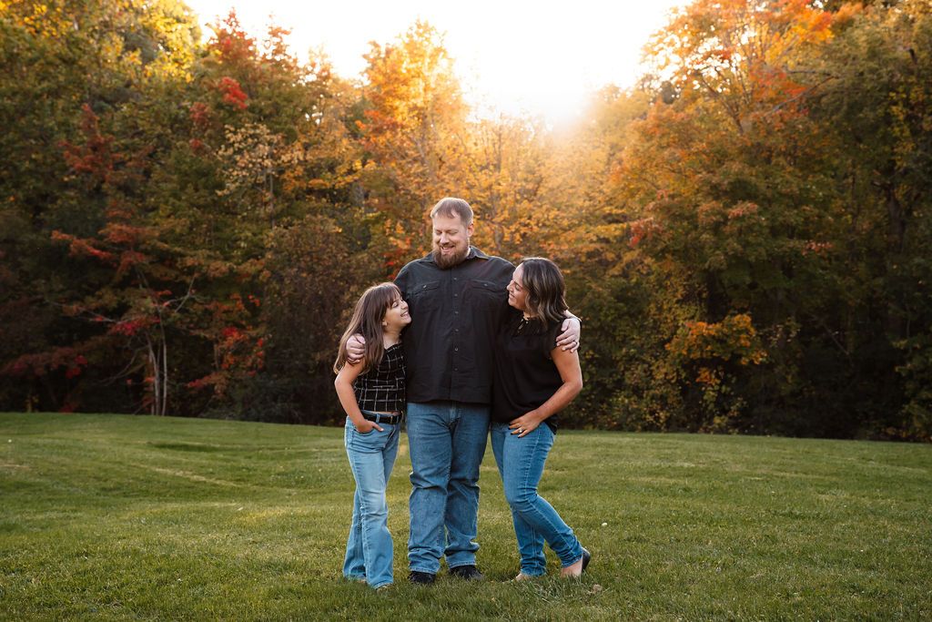 Family of three posing outdoors during golden hour with vibrant fall foliage in background on green lawn.