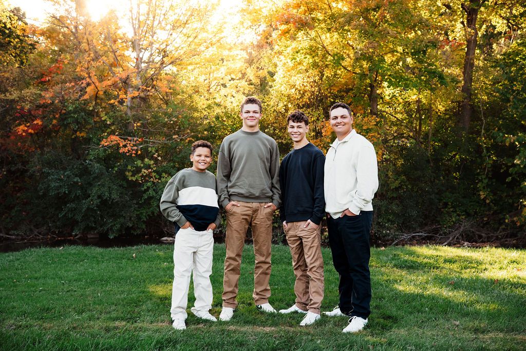 Four males posing together on a green lawn surrounded by vibrant autumn foliage during golden hour.