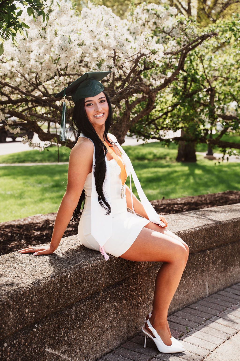 Young woman in white dress and green graduation cap sits on stone wall near blooming white trees.