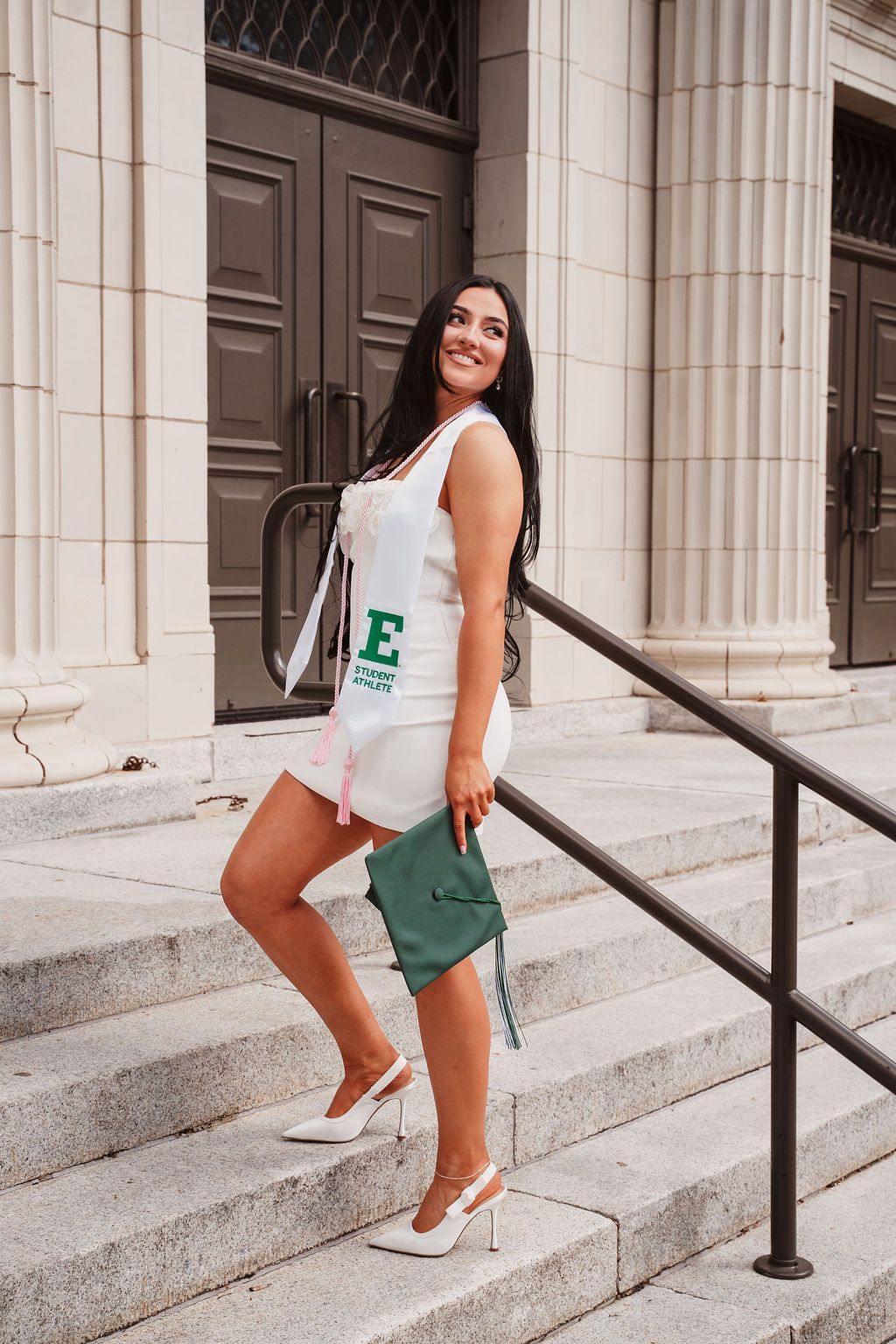 Smiling female graduate in white dress holding green cap on university steps wearing white heels.