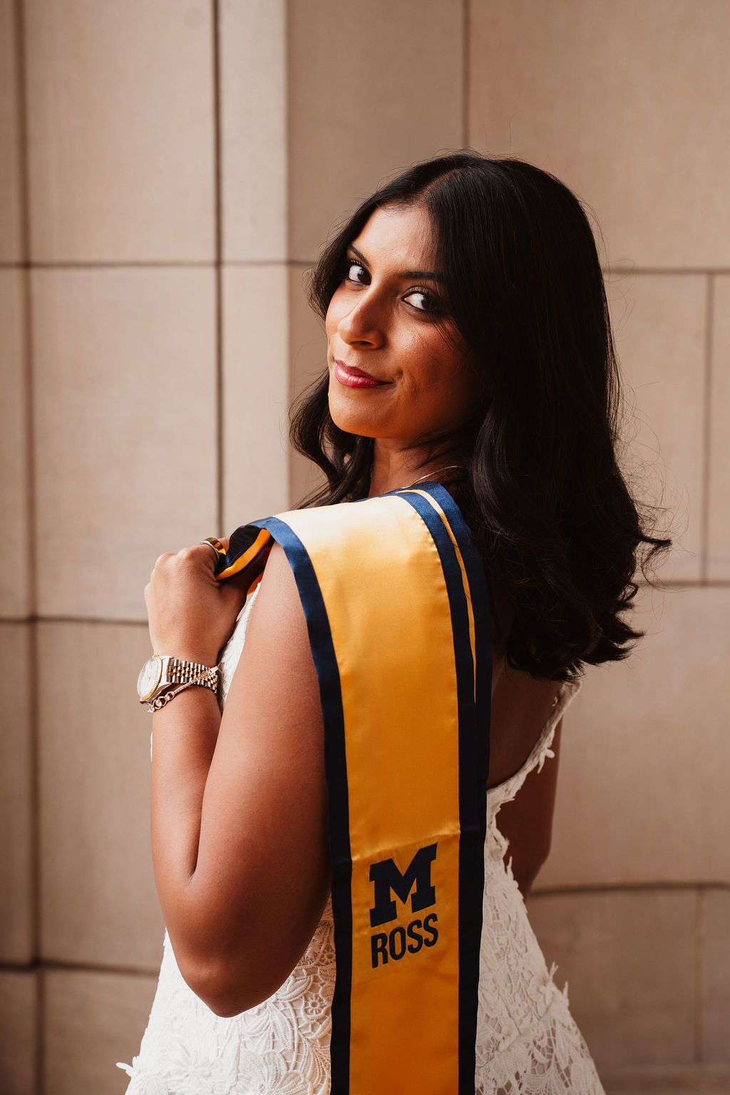 Close-up of smiling graduate in white dress holding University of Michigan Ross School gold sash over shoulder.