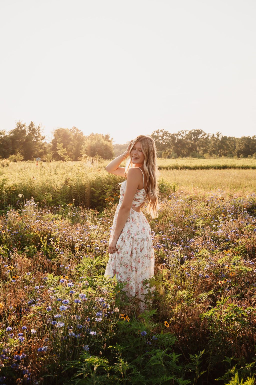 Young woman in floral dress standing in wildflower field during golden hour sunset, smiling over shoulder.
