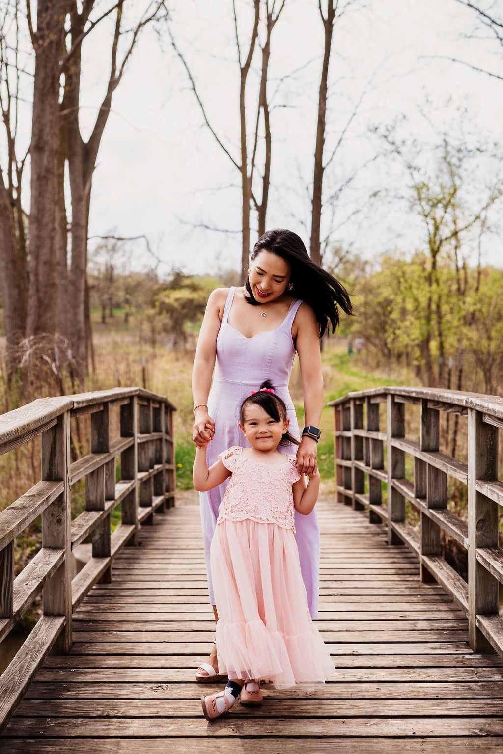 Two people in pink dresses walk together on a wooden bridge surrounded by trees in a natural setting.