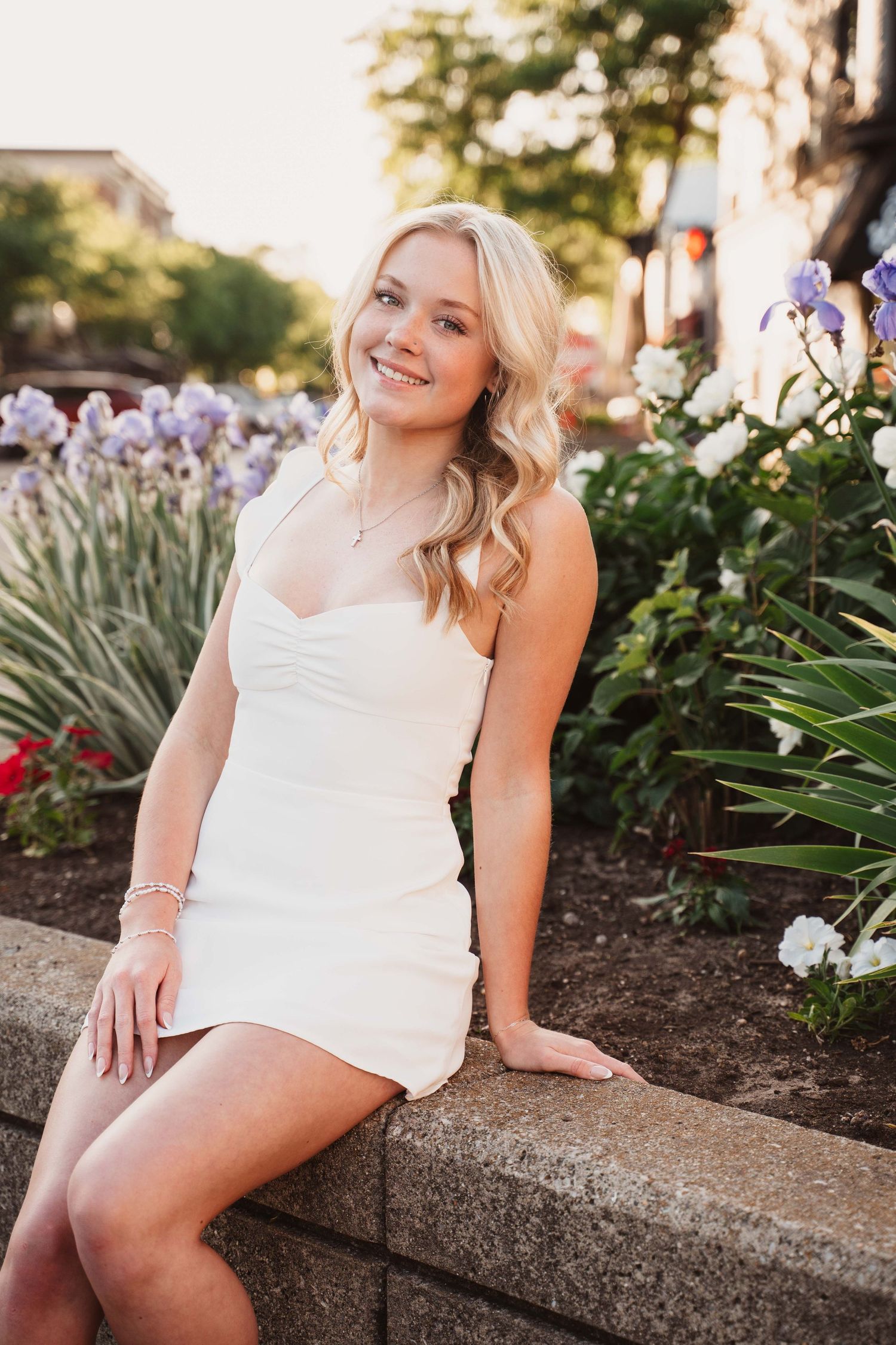 Young blonde woman in white mini dress sitting on stone wall surrounded by colorful flowers, smiling outdoors.