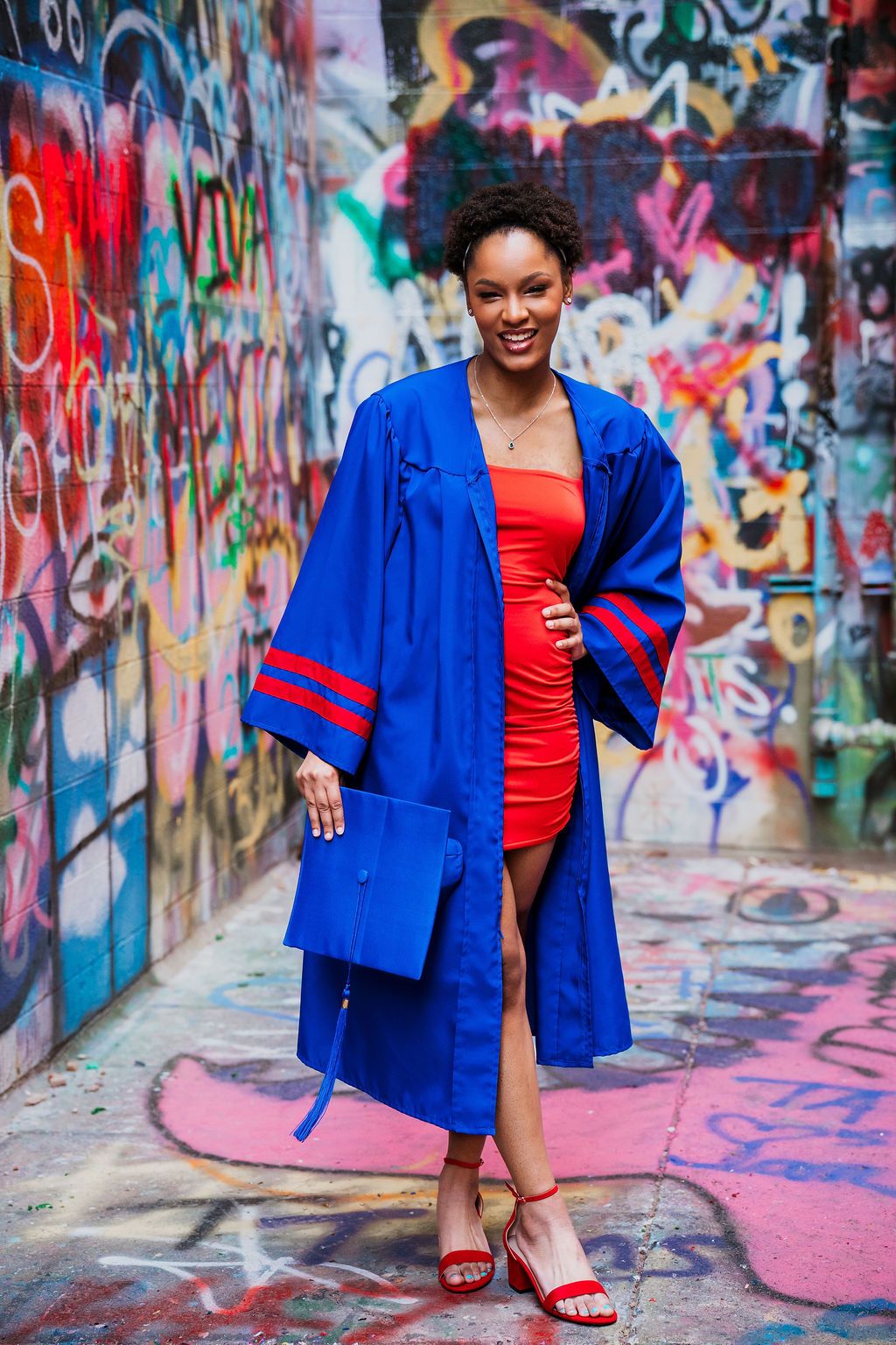 Graduate posing in front of colorful graffiti wall wearing bright blue graduation gown and red dress.