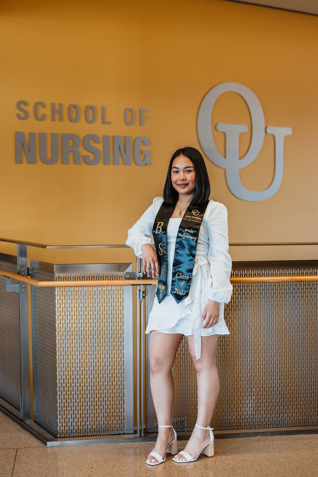 Graduate poses in front of School of Nursing signage wearing white dress and graduation gown.