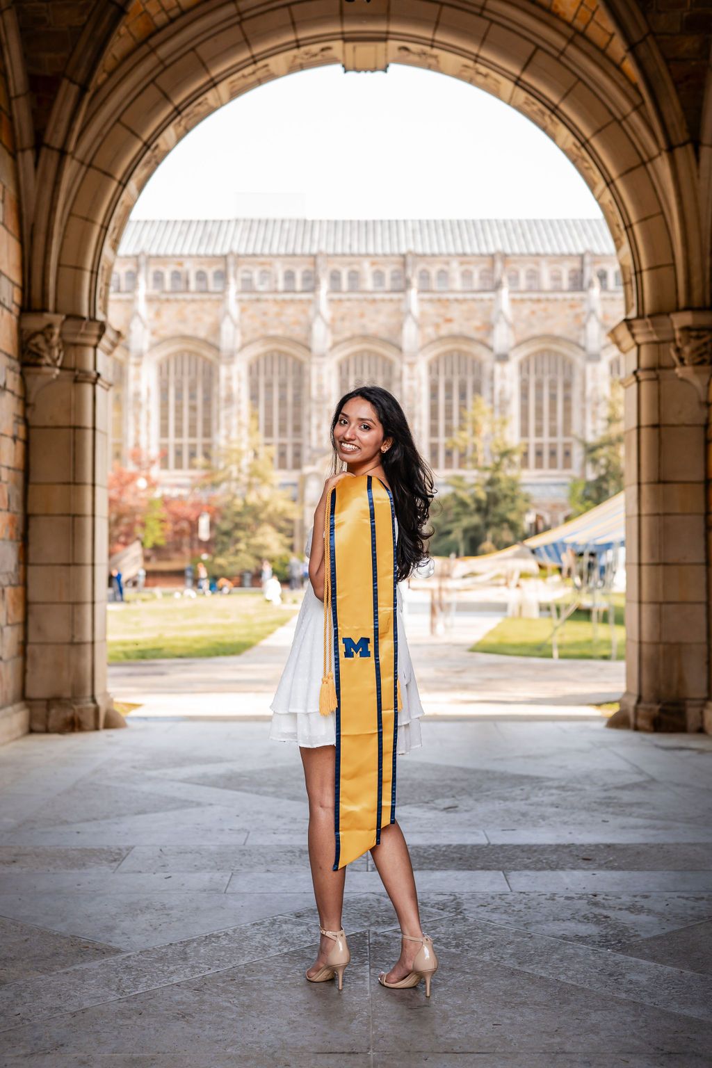 Graduate wearing yellow stole poses in stone archway with university architecture visible in background.
