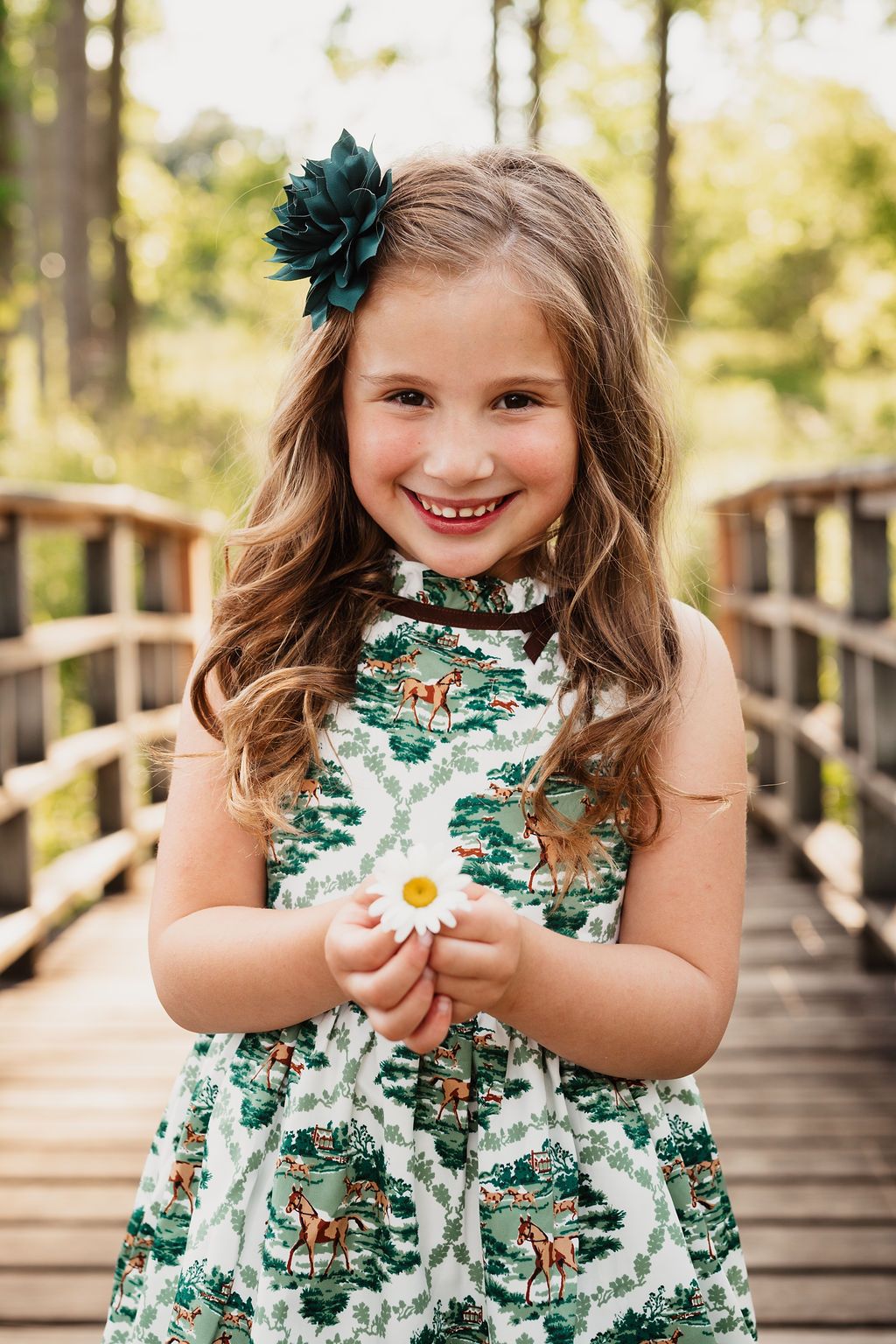 A child in a floral green and white dress holds a daisy while standing on a wooden bridge in a forest setting.