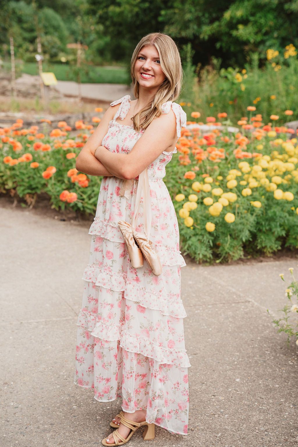 A person in a floral tiered maxi dress stands smiling on a garden path with orange and yellow flowers blooming behind.