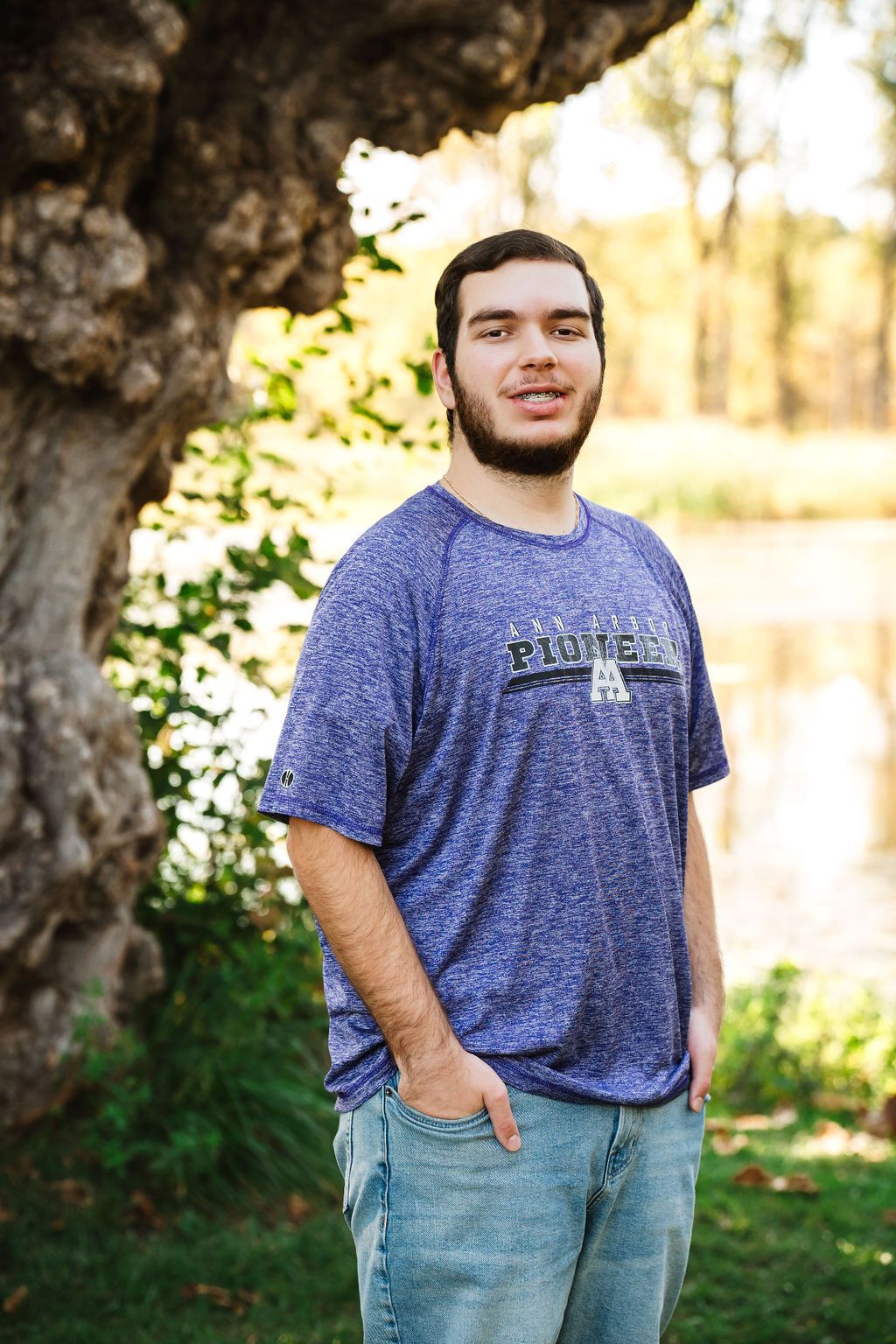 Young man in blue Pioneer t-shirt smiling outdoors near a large tree with green foliage and pond in background.